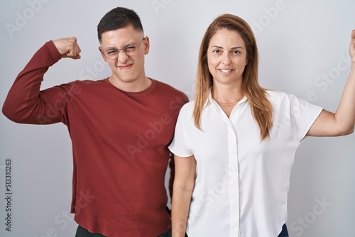 Mother and son standing together over isolated background strong person showing arm muscle, confident and proud of power