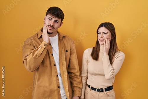 Young hispanic couple standing over yellow background touching mouth with hand with painful expression because of toothache or dental illness on teeth. dentist