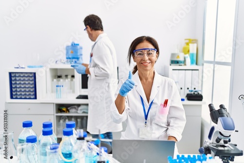 Middle age woman working at scientist laboratory smiling happy and positive  thumb up doing excellent and approval sign
