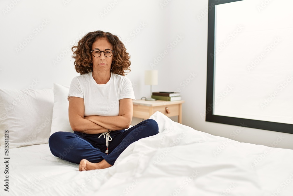 Middle age hispanic woman sitting on the bed at home skeptic and nervous, disapproving expression on face with crossed arms. negative person.