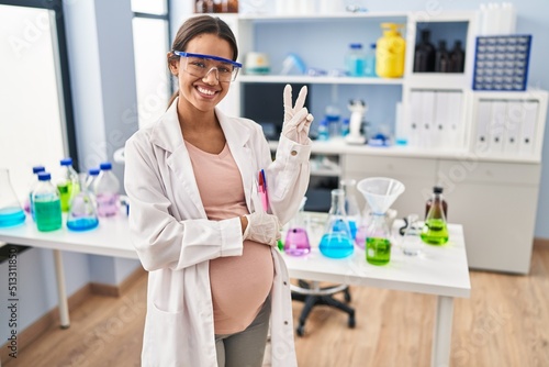 Young pregnant woman working at scientist laboratory smiling with happy face winking at the camera doing victory sign. number two.