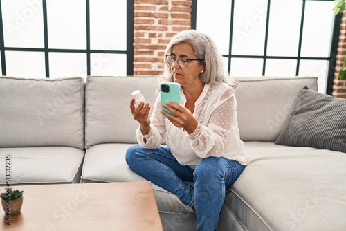 Middle age woman using smartphone holding pills at home