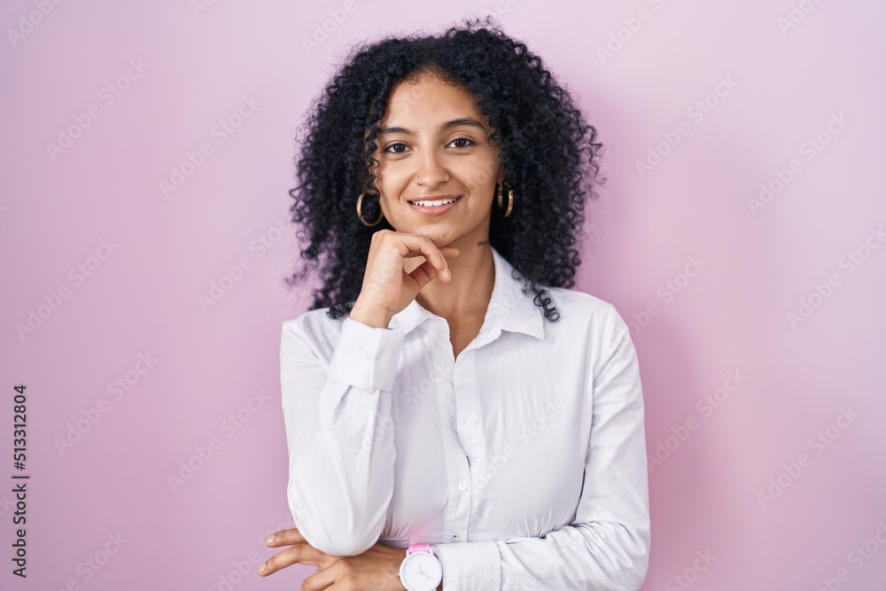 Hispanic woman with curly hair standing over pink background with hand on chin thinking about question, pensive expression. smiling and thoughtful face. doubt concept. Hispanic woman with curly hair standing over pink background with hand on chin thinking about question, pensive expression. smiling and thoughtful face. doubt concept.