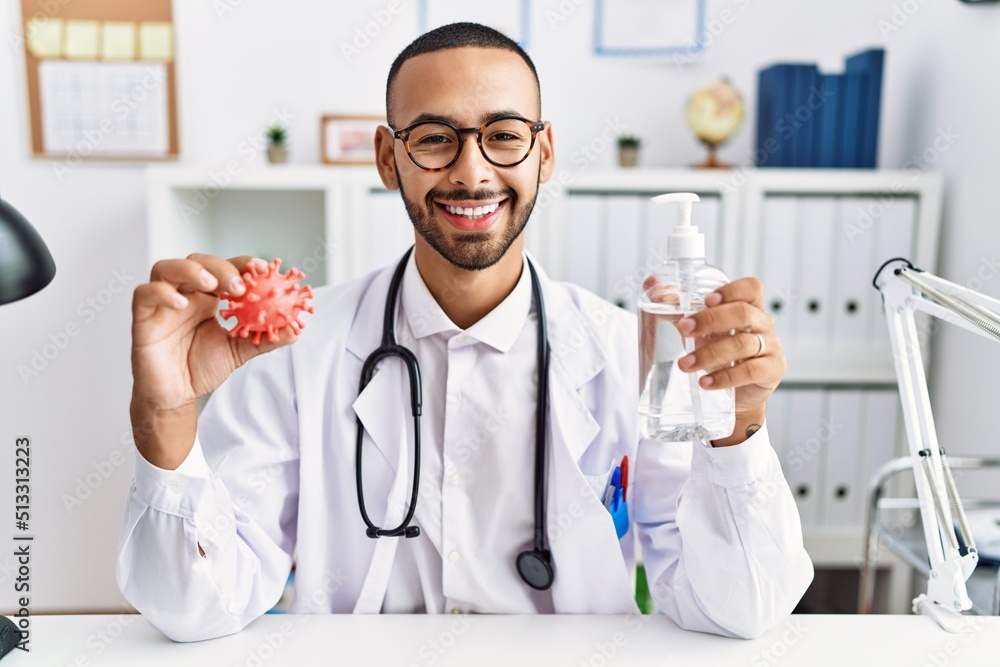 African american doctor man holding virus toy an sanitizer gel smiling with a happy and cool smile on face. showing teeth. African american doctor man holding virus toy an sanitizer gel smiling with a happy and cool smile on face. showing teeth.