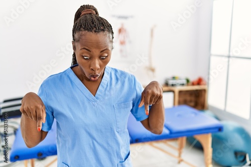 Black woman with braids working at pain recovery clinic pointing down with fingers showing advertisement, surprised face and open mouth