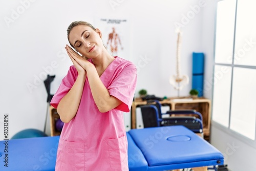 Young blonde woman working at pain recovery clinic sleeping tired dreaming and posing with hands together while smiling with closed eyes.