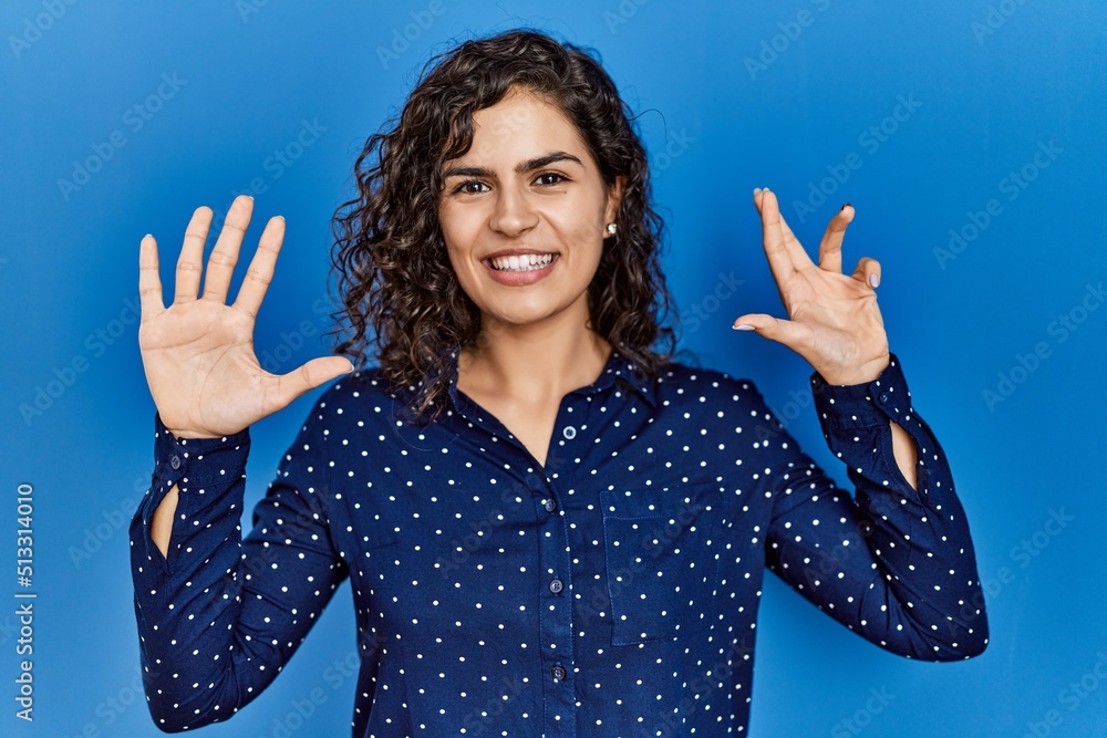Young brunette woman with curly hair wearing casual clothes over blue background showing and pointing up with fingers number nine while smiling confident and happy. Young brunette woman with curly hair wearing casual clothes over blue background showing and pointing up with fingers number nine while smiling confident and happy.