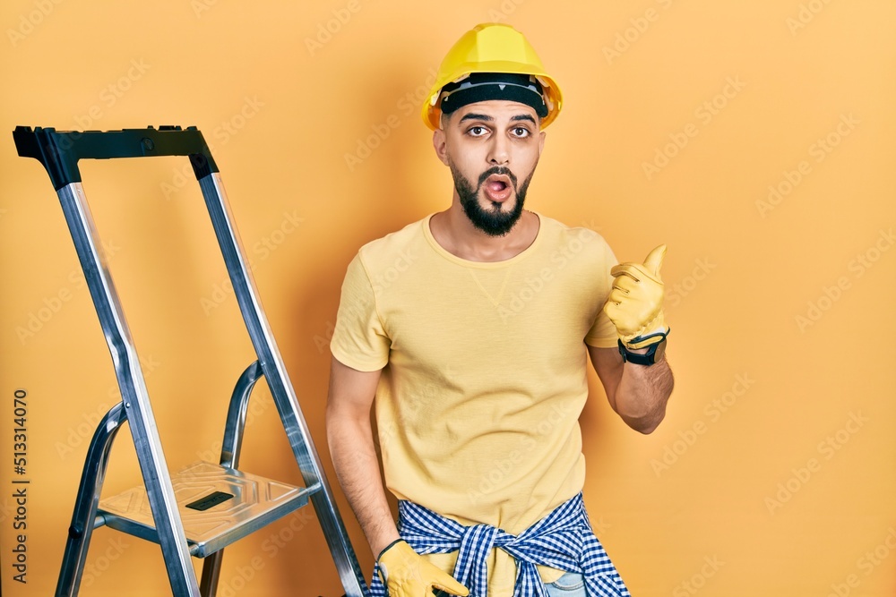 Handsome man with beard by construction stairs wearing hardhat surprised pointing with hand finger to the side, open mouth amazed expression. Handsome man with beard by construction stairs wearing hardhat surprised pointing with hand finger to the side, open mouth amazed expression.