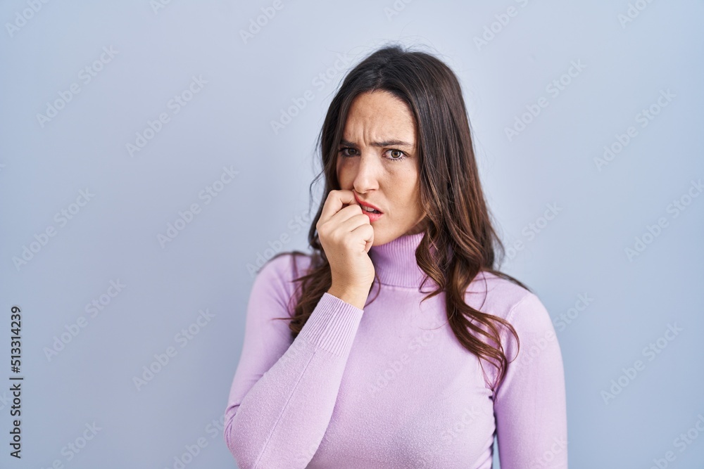 Young brunette woman standing over blue background looking stressed and nervous with hands on mouth biting nails. anxiety problem. Young brunette woman standing over blue background looking stressed and nervous with hands on mouth biting nails. anxiety problem.