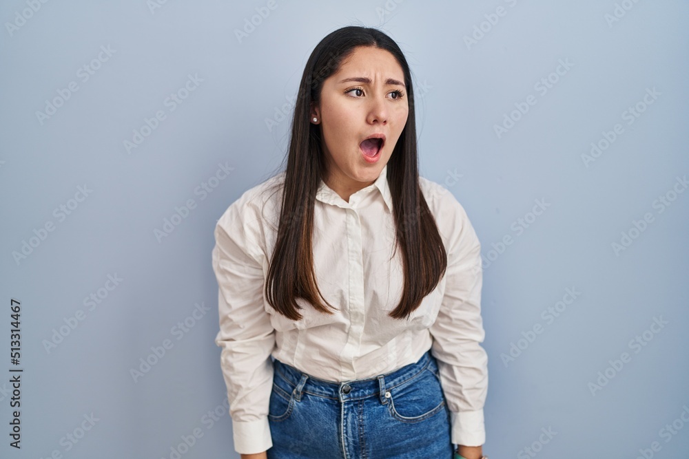 Young latin woman standing over blue background angry and mad screaming frustrated and furious, shouting with anger. rage and aggressive concept. Young latin woman standing over blue background angry and mad screaming frustrated and furious, shouting with anger. rage and aggressive concept.