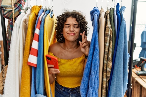Young hispanic woman searching clothes on clothing rack using smartphone gesturing finger crossed smiling with hope and eyes closed. luck and superstitious concept.