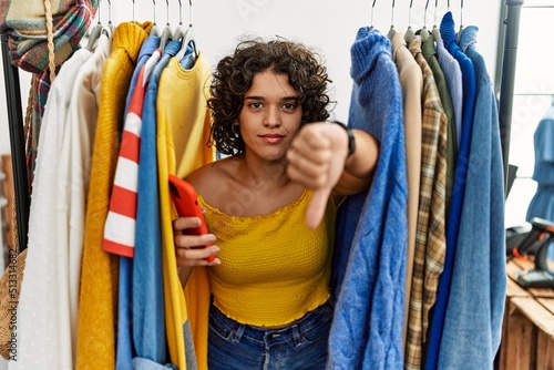 Young hispanic woman searching clothes on clothing rack using smartphone looking unhappy and angry showing rejection and negative with thumbs down gesture. bad expression.