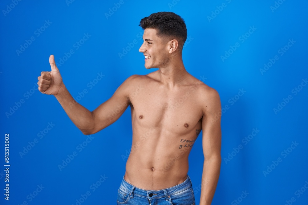 Young hispanic man standing shirtless over blue background looking proud, smiling doing thumbs up gesture to the side