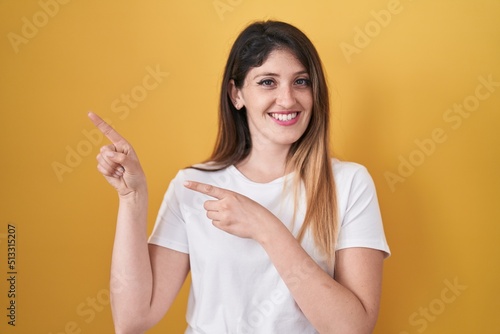 Young brunette woman standing over yellow background smiling and looking at the camera pointing with two hands and fingers to the side.