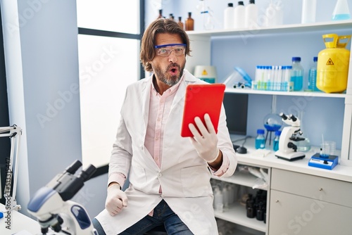 Handsome middle age man working at scientist laboratory doing video call scared and amazed with open mouth for surprise, disbelief face