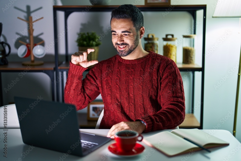 Young hispanic man with beard using computer laptop at night at home smiling and confident gesturing with hand doing small size sign with fingers looking and the camera. measure concept. Young hispanic man with beard using computer laptop at night at home smiling and confident gesturing with hand doing small size sign with fingers looking and the camera. measure concept.