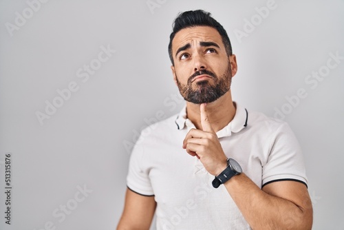Young hispanic man with beard wearing casual clothes over white background thinking concentrated about doubt with finger on chin and looking up wondering