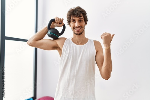Young hispanic man wearing sportswear using dumbbells pointing thumb up to the side smiling happy with open mouth © Krakenimages.com
