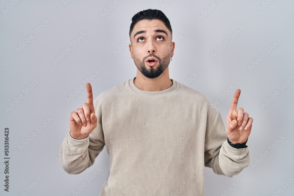 Young handsome man standing over isolated background amazed and surprised looking up and pointing with fingers and raised arms. Young handsome man standing over isolated background amazed and surprised looking up and pointing with fingers and raised arms.