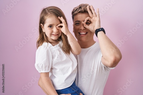 Young father hugging daughter over pink background smiling happy doing ok sign with hand on eye looking through fingers