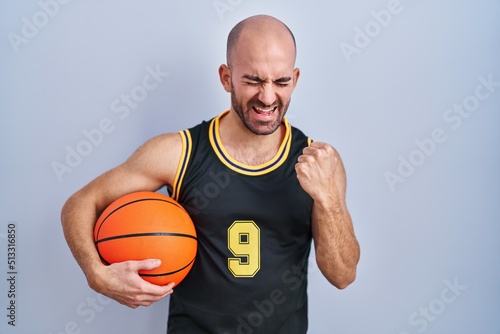 Young bald man with beard wearing basketball uniform holding ball very happy and excited doing winner gesture with arms raised, smiling and screaming for success. celebration concept.