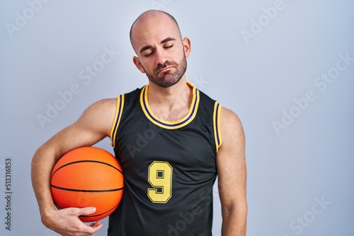 Young bald man with beard wearing basketball uniform holding ball looking sleepy and tired, exhausted for fatigue and hangover, lazy eyes in the morning.