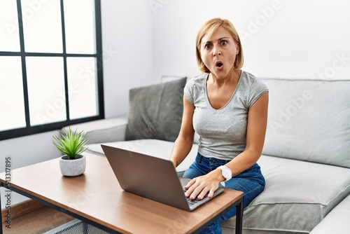Middle age blonde woman using laptop at home in shock face  looking skeptical and sarcastic  surprised with open mouth