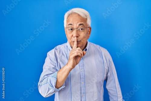 Hispanic senior man wearing glasses asking to be quiet with finger on lips. silence and secret concept.