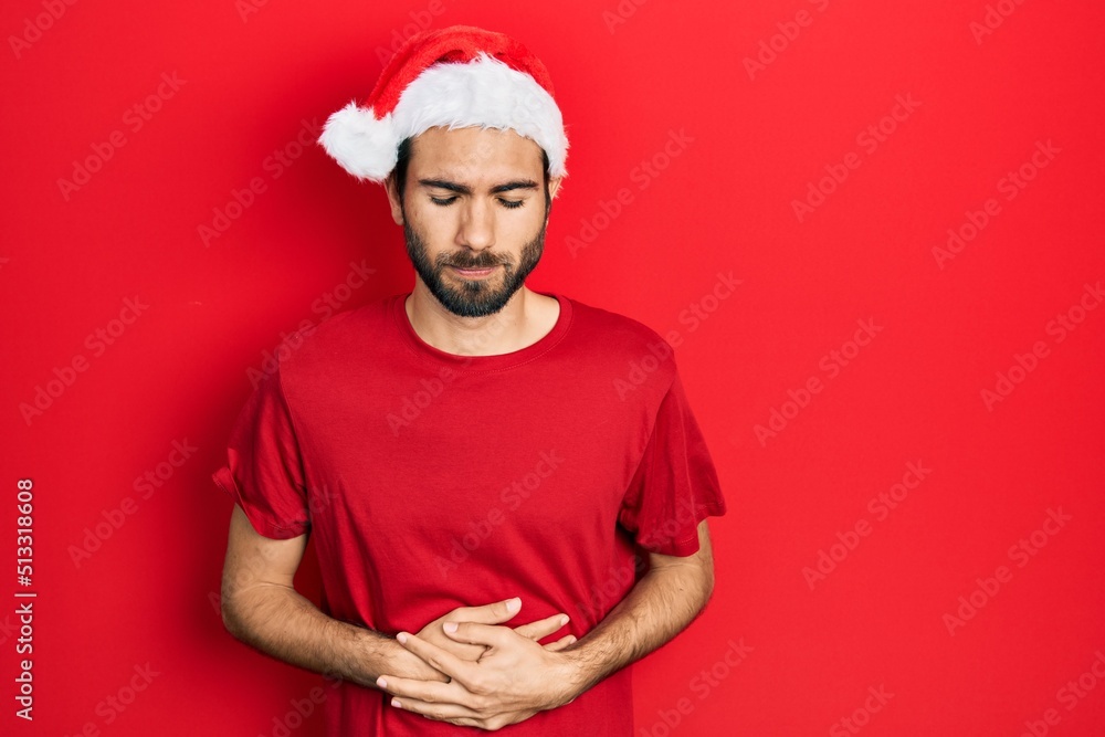 Young hispanic man wearing christmas hat with hand on stomach because indigestion, painful illness feeling unwell. ache concept.