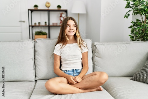 Young brunette teenager sitting on the sofa at home looking away to side with smile on face, natural expression. laughing confident.