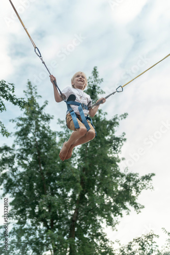 Happy blond boy hangs on slings, jumps high on trampoline in an amusement park. Schoolboy is having fun.