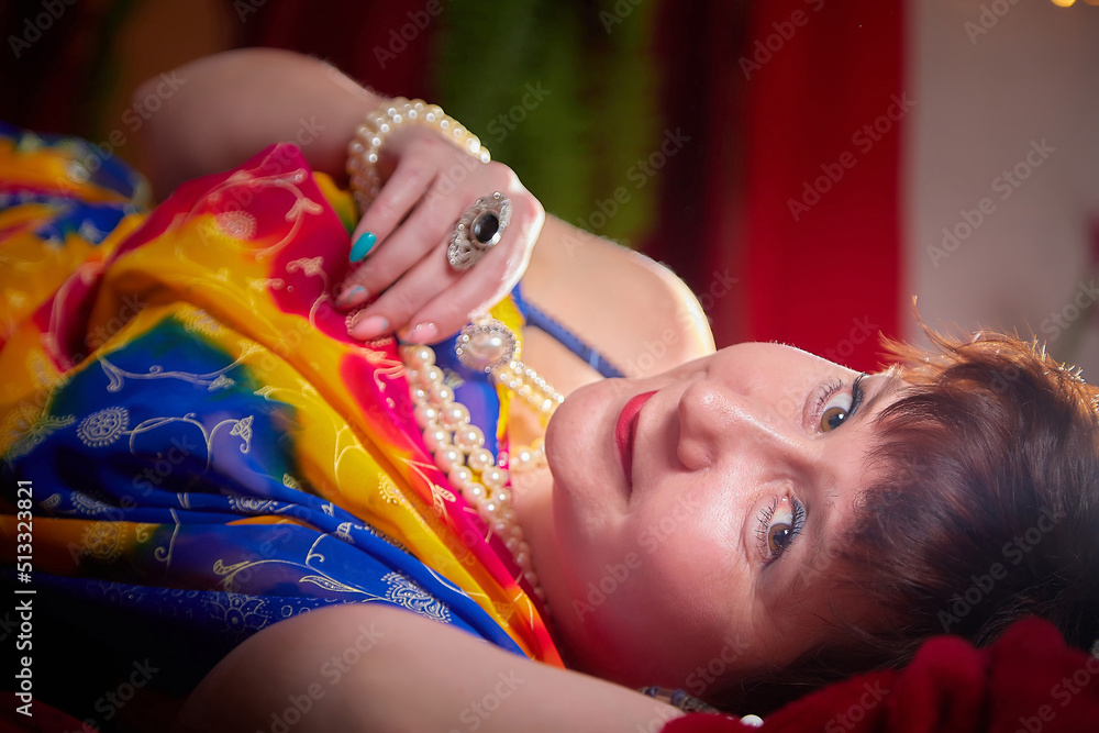 Naklejka premium A beautiful European girl with short hair looking like an Arab woman in a red room in a harem. Photo shoot of an oriental style odalisque. A model poses in a sari