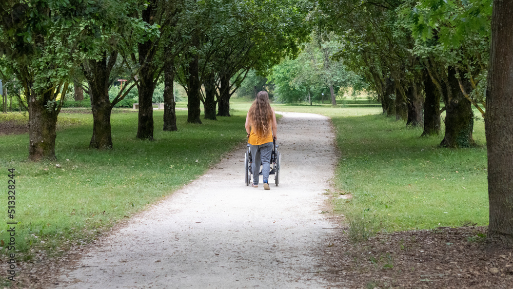 Fototapeta premium Young woman with long hair pushing a wheelchair through the middle of a tree-lined driveway in a park, rear view