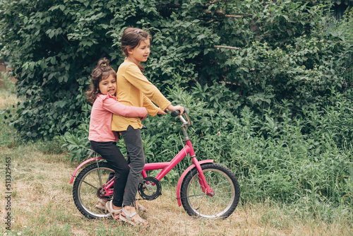 Two caucasian girls friends rides a bike through the countryside, care and sisterly love, Ukrainian children and village
