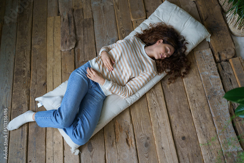 Beautiful young calm Italian woman lying on mattress on wooden floor at home, peaceful Hispanic female sleeping, napping, getting some rest during day, top view, full length image. Napping Meditation © DimaBerlin