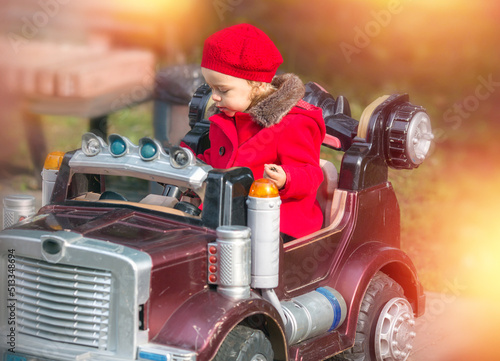 Fototapeta Naklejka Na Ścianę i Meble -  Happy caucasian little girl wearing a red coat and beret in the park while sitting sitting in a four-wheeled children's car. Childhood. Autumn Park. Entertainment concept