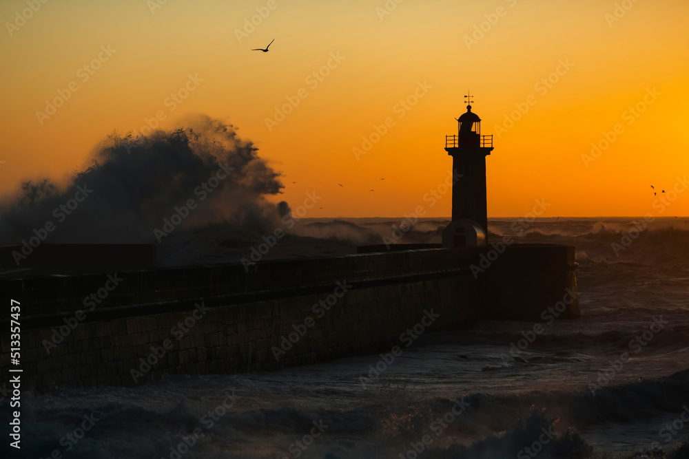 The silhouette of a lighthouse being washed by a huge wave during sunset.
