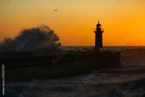 The silhouette of a lighthouse being washed by a huge wave during sunset.
