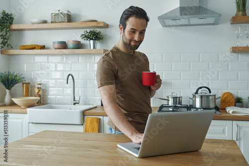 Handsome young man using laptop and enjoying coffee while standing at the kitchen island