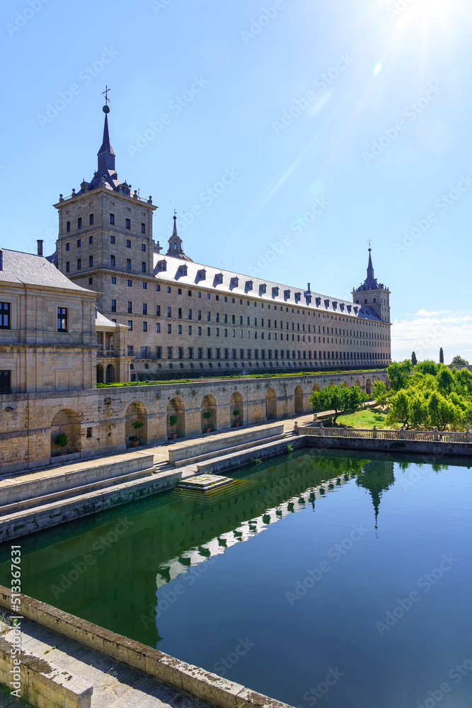 Monastery of El Escorial with water pond, gardens and flashes of sun in the blue sky.