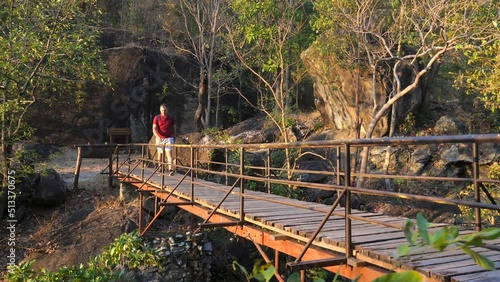 Wallpaper Mural Adult man with backpack walk on pedestrian bridge in woods during sunset time, covers eyes with hand. Happy male tourist go hiking or trekking on wooden path in natural park. Tourism, travel concept. Torontodigital.ca