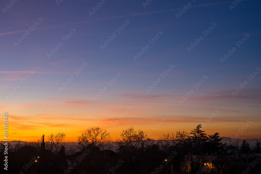 Colourful sunset with clear sky and some clouds from the city, with the silhouettes of trees and some buildings in front of the mountains on the horizon