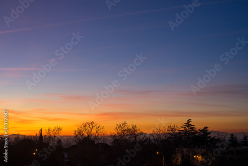 Colourful sunset with clear sky and some clouds from the city, with the silhouettes of trees and some buildings in front of the mountains on the horizon © Adrian Zarzuelo