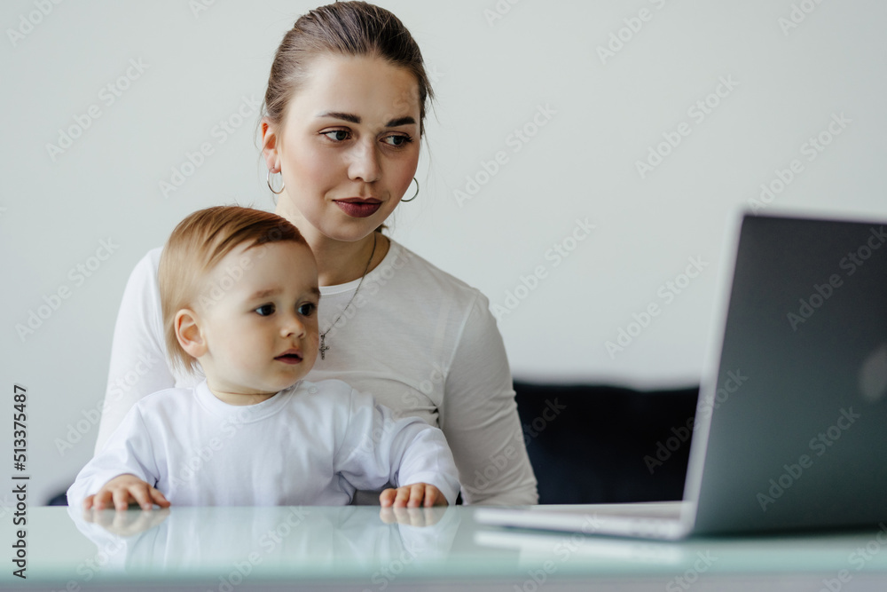 Young single mother mom with toddler newborn baby infant son using laptop, searching web, watching cartoons together. Working from home on maternity leave, freelancer, looking for kids goods Young single mother mom with toddler newborn baby infant son using laptop, searching web, watching cartoons together. Working from home on maternity leave, freelancer, looking for kids goods