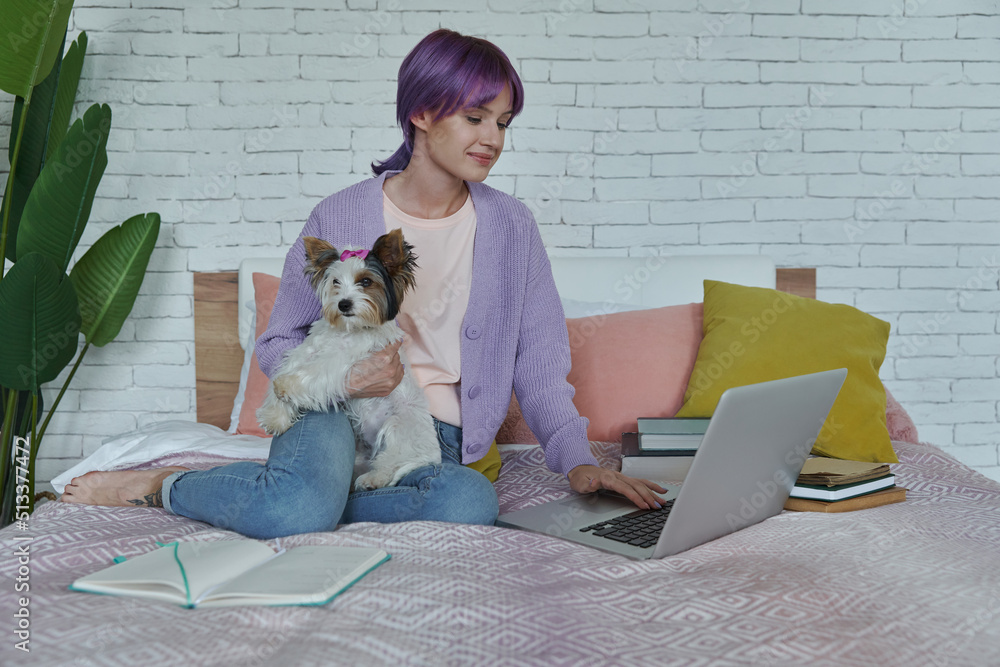 Confident teenage girl using laptop while sitting on bed with her dog