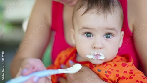 Cute baby sitting on mom's laps being fed from spoon. Lovely kid taking food and wrinkling up. Close up.