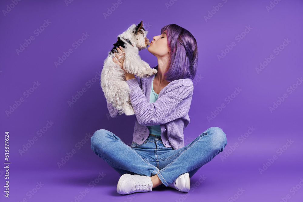 Playful young woman carrying little dog while sitting against purple background Playful young woman carrying little dog while sitting against purple background