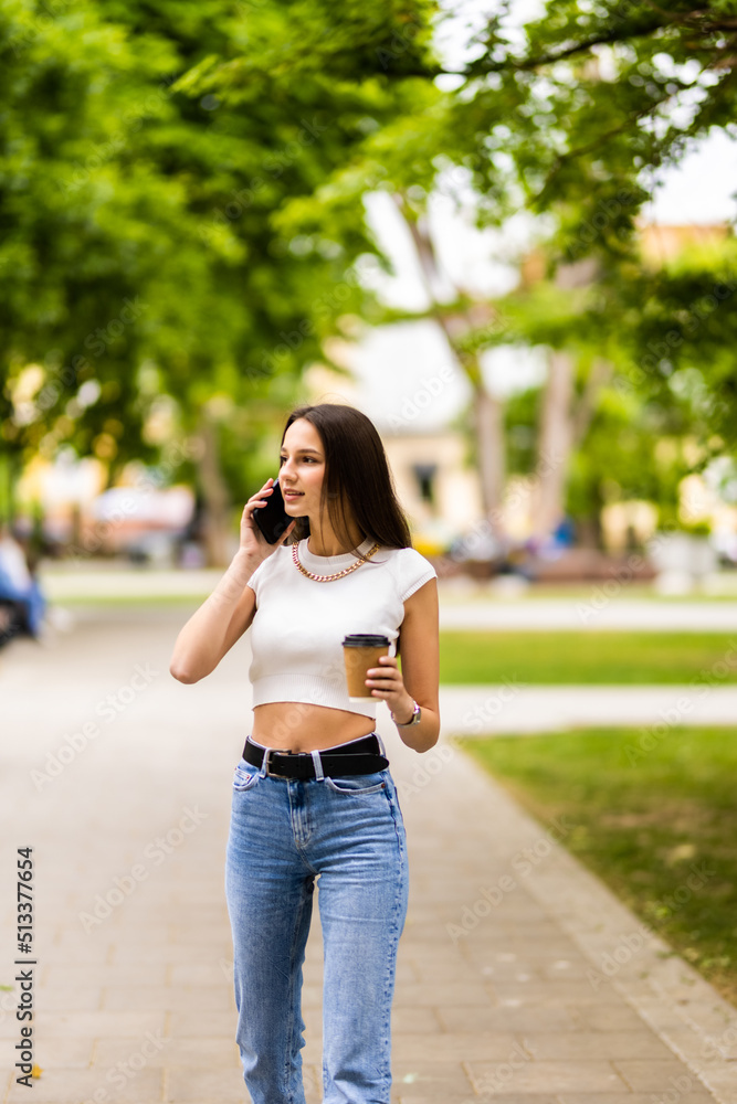 Caucasian business woman speaking by phone holding coffee to go. A successful European woman, talking on the phone, standing on modern office building background