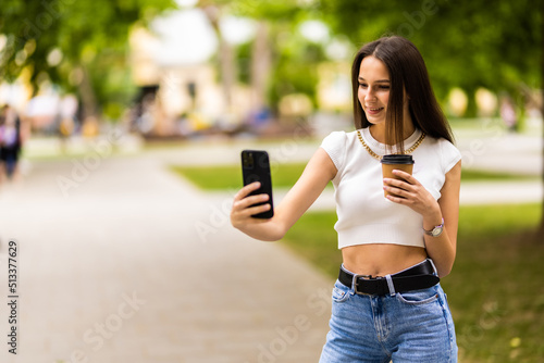 Happy woman, attractive girl holding takeaway cup, walking in city with smartphone in hand, looking aside. Girl going somewhere on street, drinking coffee and chat on mobile phone