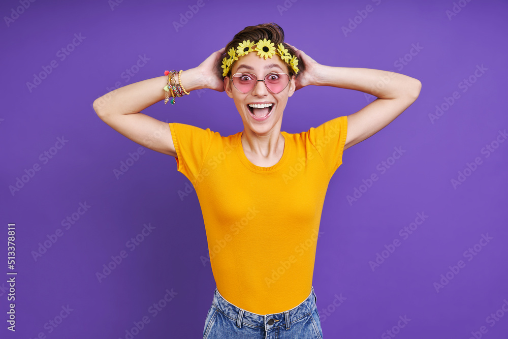 Surprised woman with floral head wreath touching head while standing against purple background